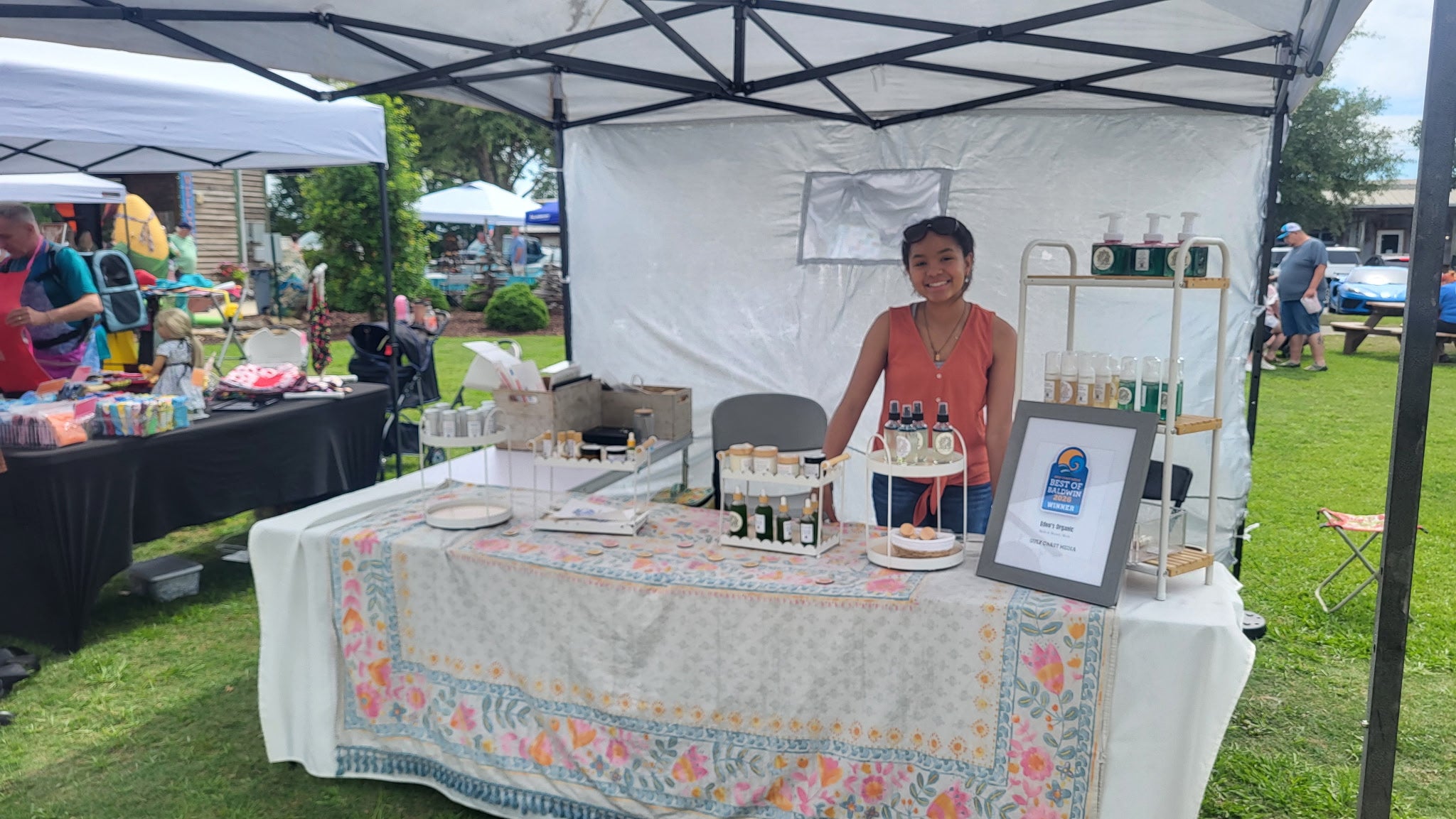 Woman standing behind a table with products under a tent at an outdoor event.