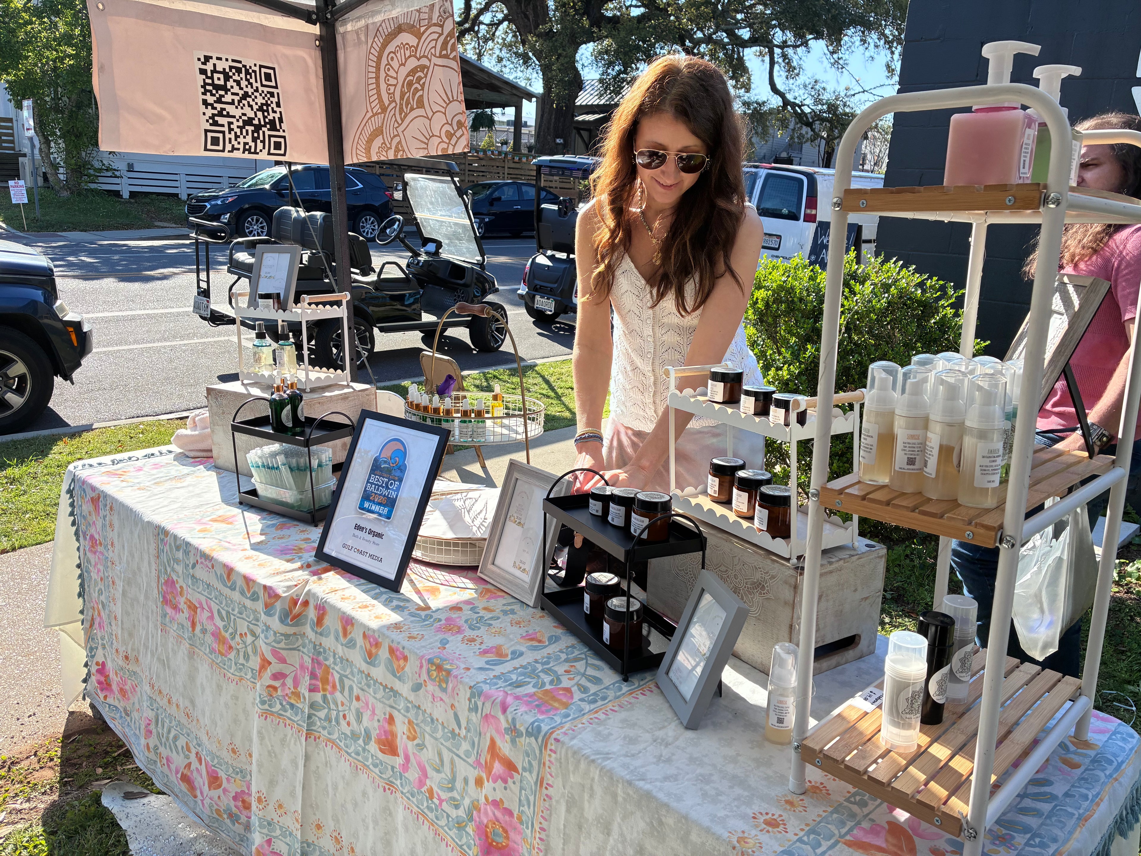 Woman at an outdoor market stall with products on a table and shelves.